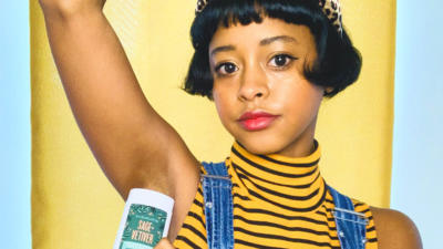 Woman with short black hair and leopard headband in front of a yellow and blue background with her arm up applying Schmidt's Sage+Vetiver Hemp Seed Oil Deodorant to her underarm